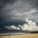 A lone figure walks along a wide golden beach beneath towering dark storm clouds, with white waves breaking along the shoreline.