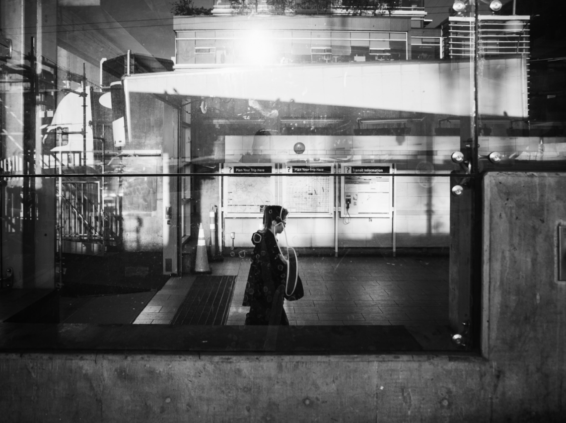 Black-and-white mobile photograph of a solitary person walking through an urban transit station, seen through reflective glass. Layered reflections, architectural structures, signage and strong bands of light create an abstract, fragmented city scene.