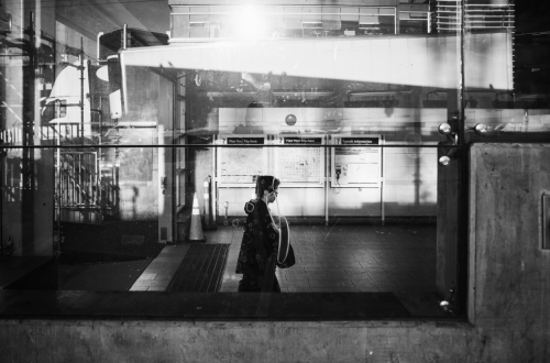 Black-and-white mobile photograph of a solitary person walking through an urban transit station, seen through reflective glass. Layered reflections, architectural structures, signage and strong bands of light create an abstract, fragmented city scene.