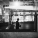 Black-and-white mobile photograph of a solitary person walking through an urban transit station, seen through reflective glass. Layered reflections, architectural structures, signage and strong bands of light create an abstract, fragmented city scene.