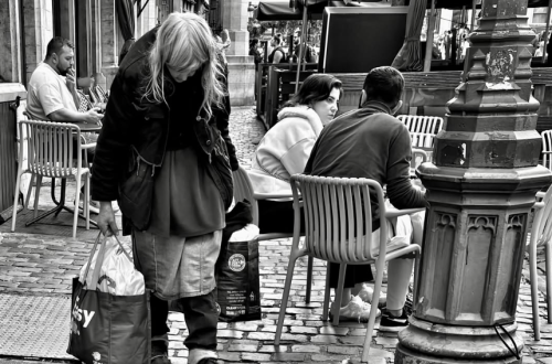 Black-and-white street photograph of a woman carrying shopping bags as she walks through a cobbled city square past an outdoor café. Seated diners, ornate architecture, awnings and a large decorative lamppost create layered depth around the solitary foreground figure.