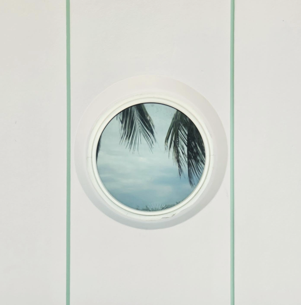 Minimalist architectural photograph featuring a circular porthole-style window set into a pale wall with two vertical mint-green lines. Palm fronds and a soft blue sky are reflected in the glass, creating a serene, almost dreamlike composition.