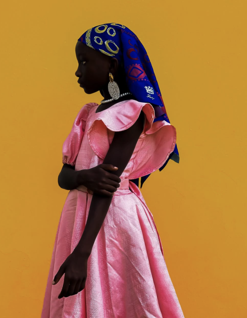 Portrait of a young girl in profile against a vibrant golden-yellow background, wearing a shimmering pink dress with sculptural ruffled sleeves, a richly patterned royal-blue headscarf, large silver earrings and a pearl necklace. Her arms are gently crossed, and her gaze is directed to the left, creating a striking composition of bold colours, contrasting textures and quiet elegance.
