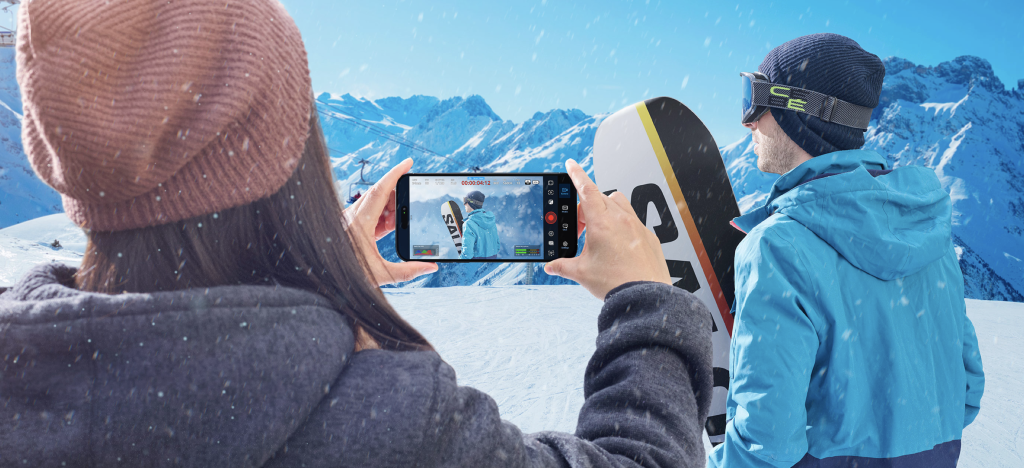 Photographer recording a snowboarder in a snowy mountain landscape using the Blackmagic Camera video app on an iPhone.