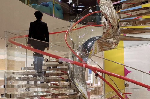 Person ascending a glass and metal staircase beside a reflective silver sculpture, surrounded by colourful architectural details and sweeping red handrails.