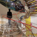 Person ascending a glass and metal staircase beside a reflective silver sculpture, surrounded by colourful architectural details and sweeping red handrails.