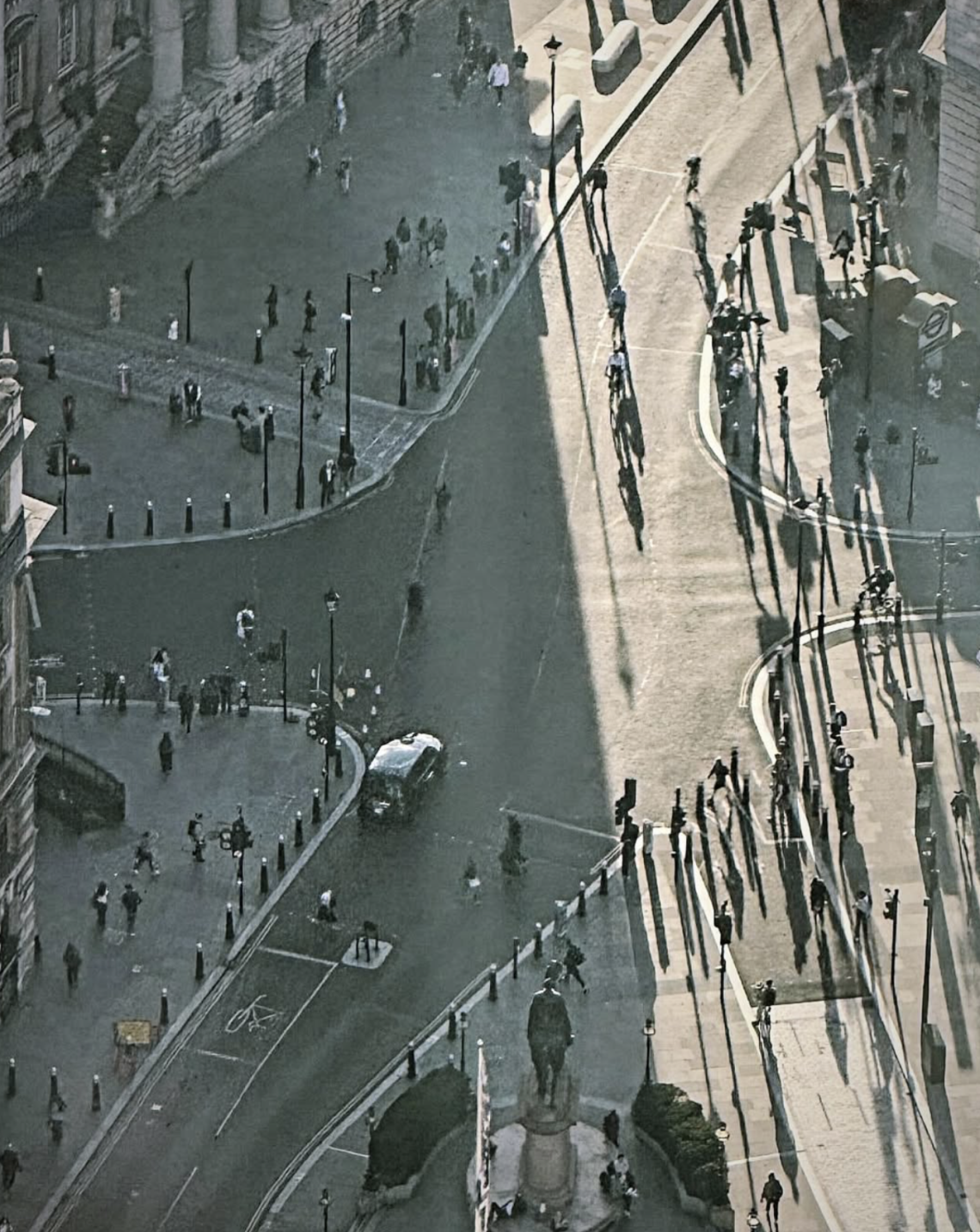 High-angle photograph looking down over a sweeping urban intersection surrounded by historic stone buildings. Pedestrians, cyclists and a single car move through the streets, while dramatic late-afternoon sunlight casts exceptionally long shadows across the pale road surface. A dark statue on a pedestal stands in the foreground, anchoring a complex composition of curved roads, architectural details and contrasting areas of light and shade.
