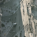 High-angle photograph looking down over a sweeping urban intersection surrounded by historic stone buildings. Pedestrians, cyclists and a single car move through the streets, while dramatic late-afternoon sunlight casts exceptionally long shadows across the pale road surface. A dark statue on a pedestal stands in the foreground, anchoring a complex composition of curved roads, architectural details and contrasting areas of light and shade.