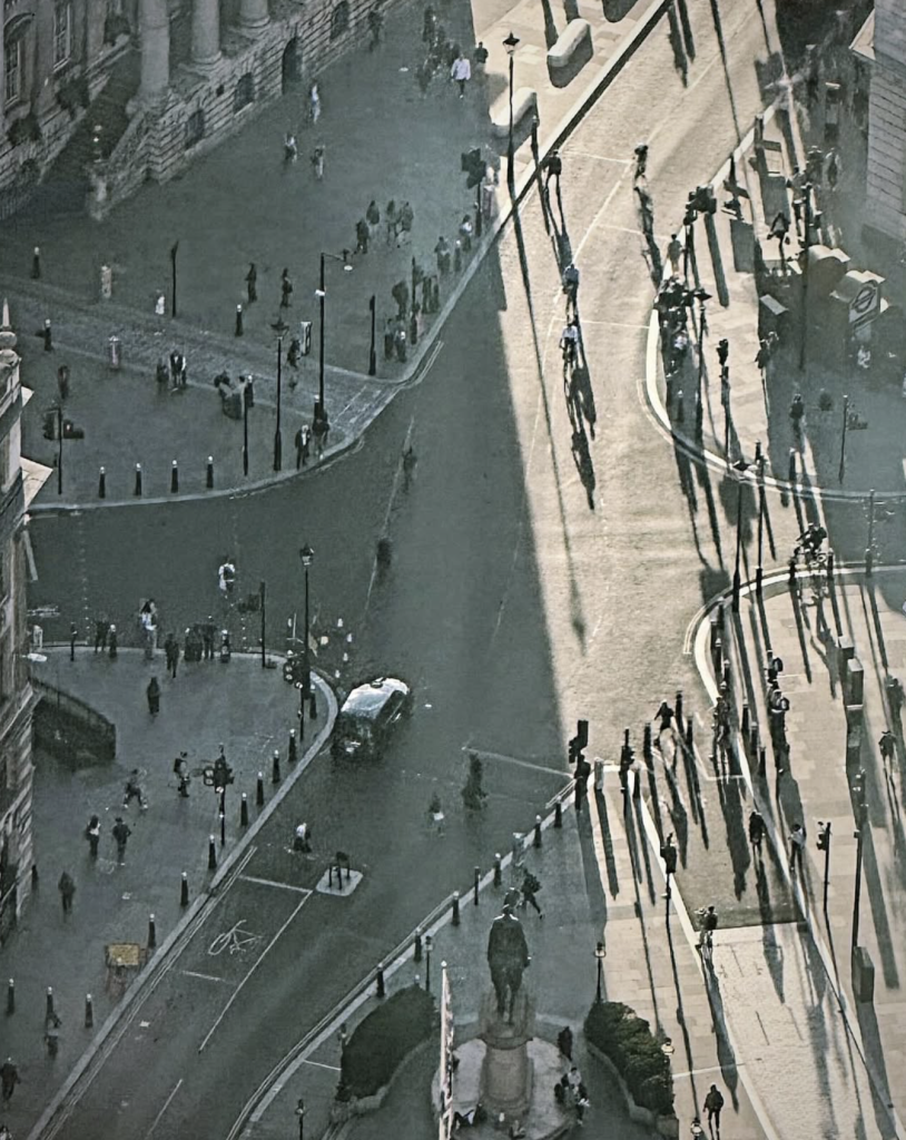High-angle photograph looking down over a sweeping urban intersection surrounded by historic stone buildings. Pedestrians, cyclists and a single car move through the streets, while dramatic late-afternoon sunlight casts exceptionally long shadows across the pale road surface. A dark statue on a pedestal stands in the foreground, anchoring a complex composition of curved roads, architectural details and contrasting areas of light and shade.