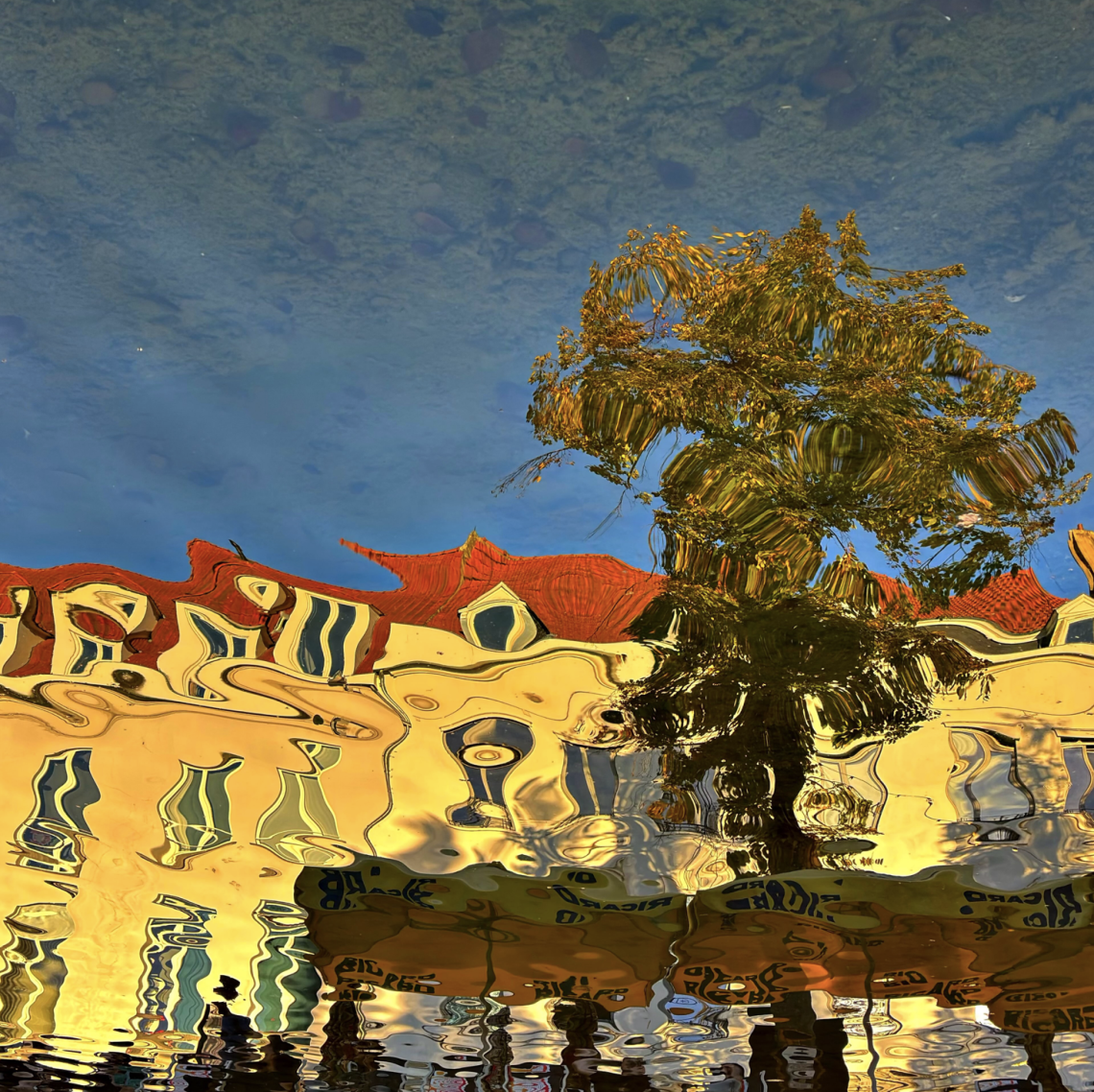 Colourful water reflection of yellow buildings with red-tiled roofs and a leafy tree, distorted by ripples into flowing, abstract shapes beneath an expanse of blue.