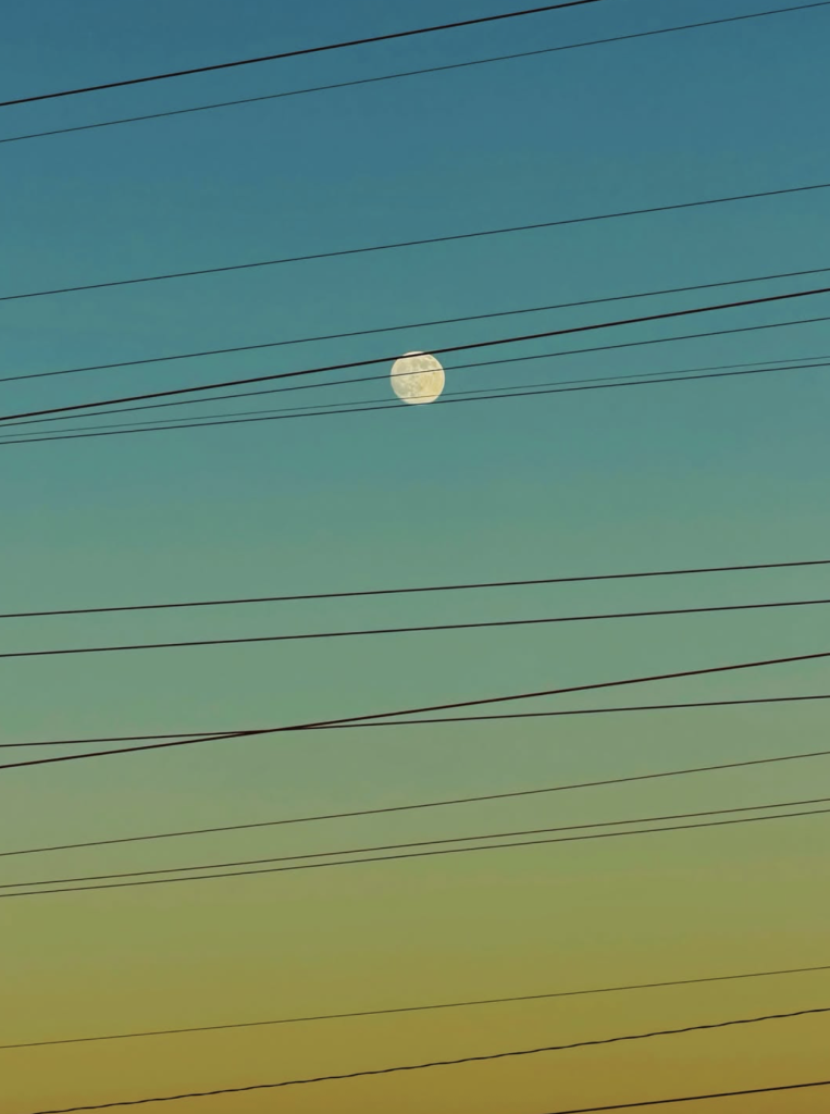 A pale, nearly full moon suspended in a turquoise evening sky, crossed by multiple dark overhead wires. The sky gradually changes from blue-green to warm yellow near the bottom of the photograph.