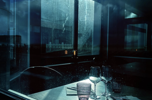Moody blue-toned photograph of an empty restaurant table with two wine glasses, drinking glasses, cutlery and folded napkins. Rain-streaked windows and reflections create a cinematic atmosphere.