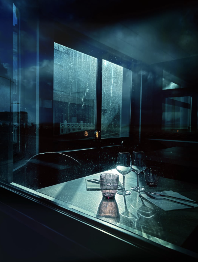 Moody blue-toned photograph of an empty restaurant table with two wine glasses, drinking glasses, cutlery and folded napkins. Rain-streaked windows and reflections create a cinematic atmosphere.