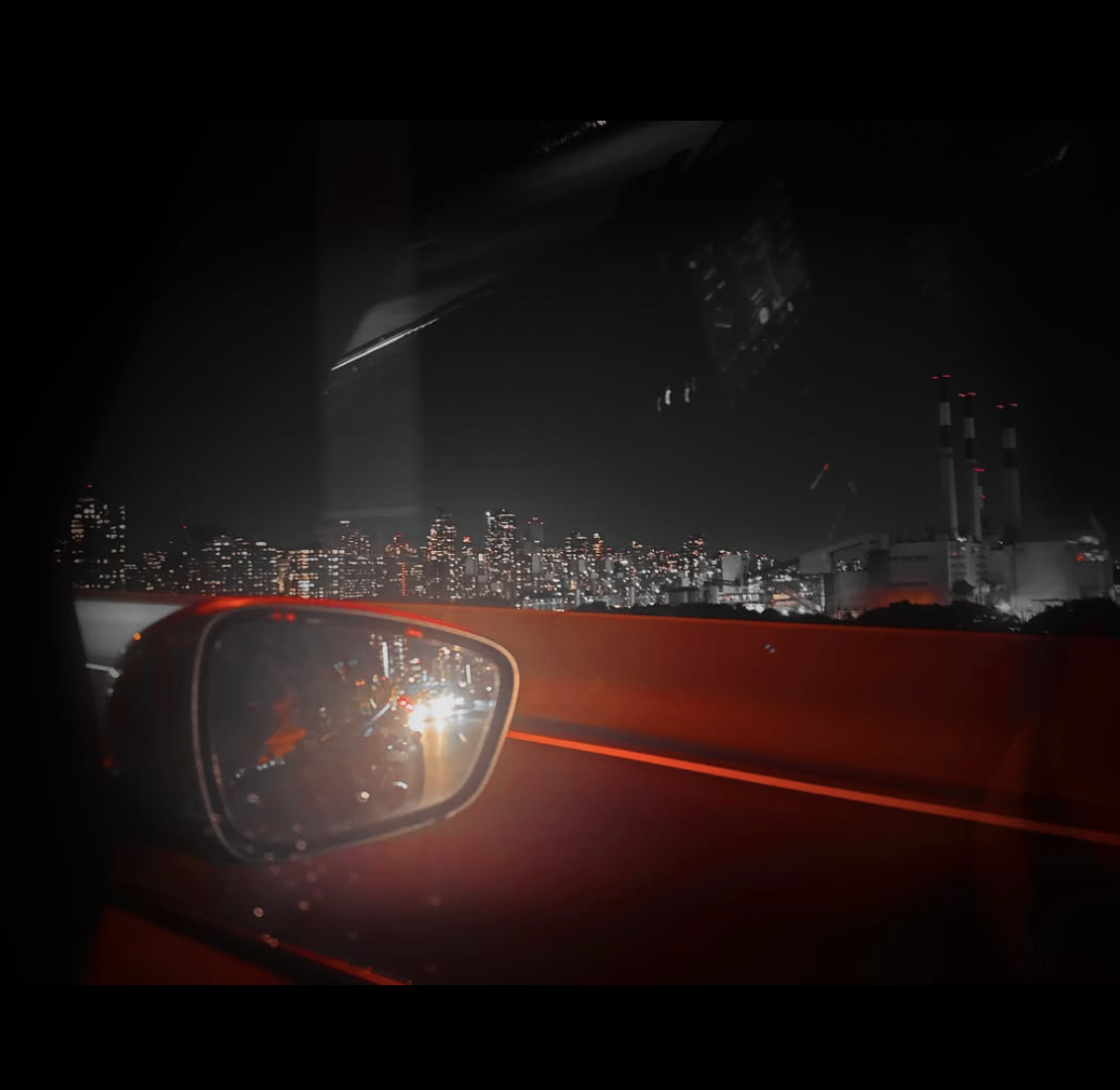 Nighttime urban photograph taken from inside a car, showing a side-view mirror reflecting approaching headlights, a red-lit road barrier and a distant illuminated city skyline.